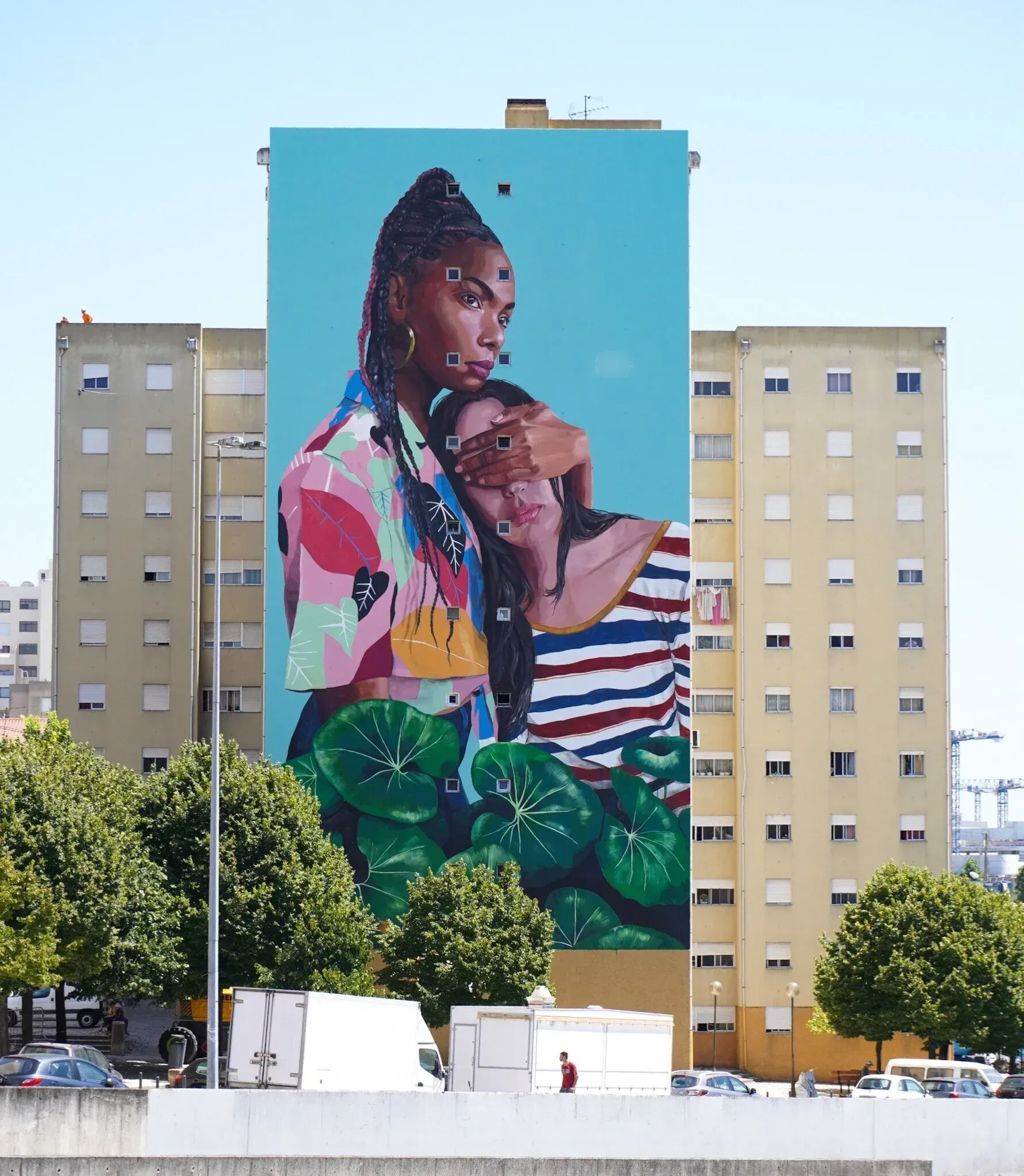 P E R S P E C T I V E S. Mural urbano que representa un abrazo protector entre dos mujeres con un fondo azul y hojas verdes. La obra transmite serenid